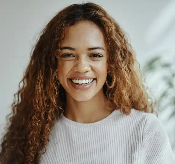 Smiling woman with curly auburn hair in casual white ribbed shirt, representing dental practice values.