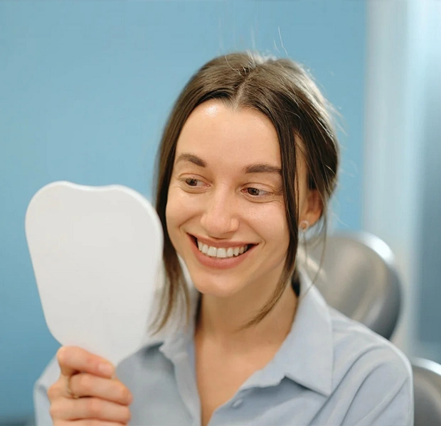 Woman smiling at a handheld mirror while sitting in a dentist's chair